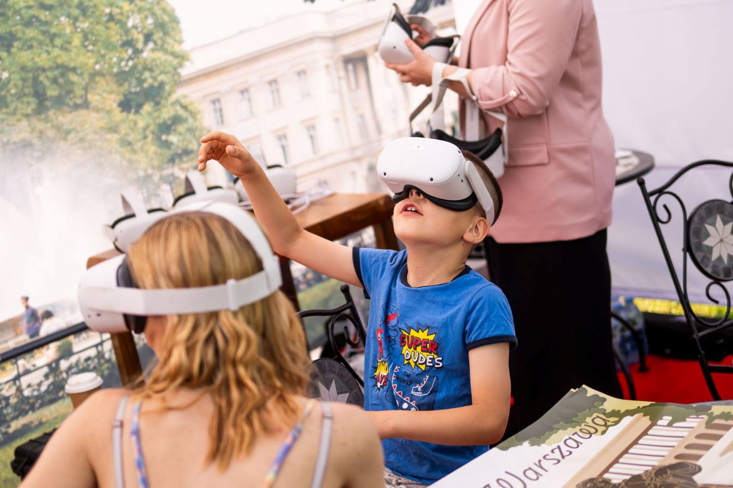 Dwójka dzieci siedząca przy stoliku. Na głowach mają nałożone okulary VR, przez które mogą oglądać multimedialny pokaz. W tle dorosła osoba trzymające takie same okulary, a za nią plakat przedstawiający Pałac Saski.