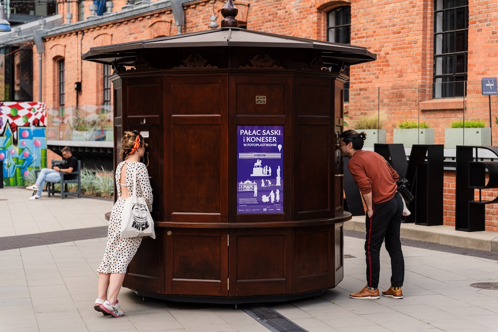 Drewniany kiosk, czyli fotoplastikon, umieszczony na dziedzińcu konesera na Pradze. Urządzenie w kształcie rotundy ma w ścianach wizjery, przez które dwie osoby zaglądają do jego środka. Za fotoplastikonem rysuje się ceglana ściana budynku.