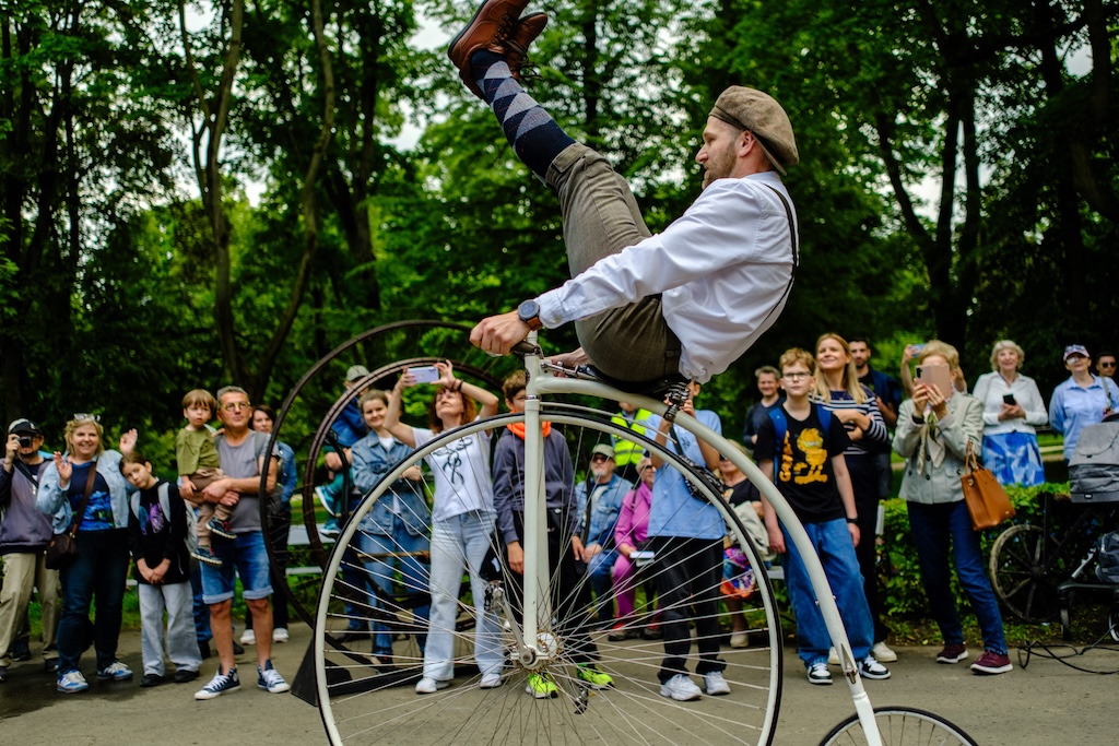 Święto Ogrodu Saskiego, czyli urodziny Ogrodu Saskiego. Mężczyzna w stroju retro, z kaszkietem i pumpami demonstruje figurę jadąc na bicyklu. Mężczyzna siedzi na siodełku tak, że nogi utrzymuje wysoko nad kierownicą, zamiast trzymać je w dole. W tle tłum ogląda pokaz.