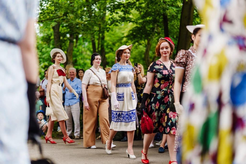Kobiety w przedwojennych strojach przechadzają się rzędem przed tłumem ludzi. Każda ma lekko przekrzywiony kapelusik i elegancki strój. Większość jest w letnich sukienkach, jedna kobieta jest w szerokich spodniach i bluzce. W tle rosną drzewa.