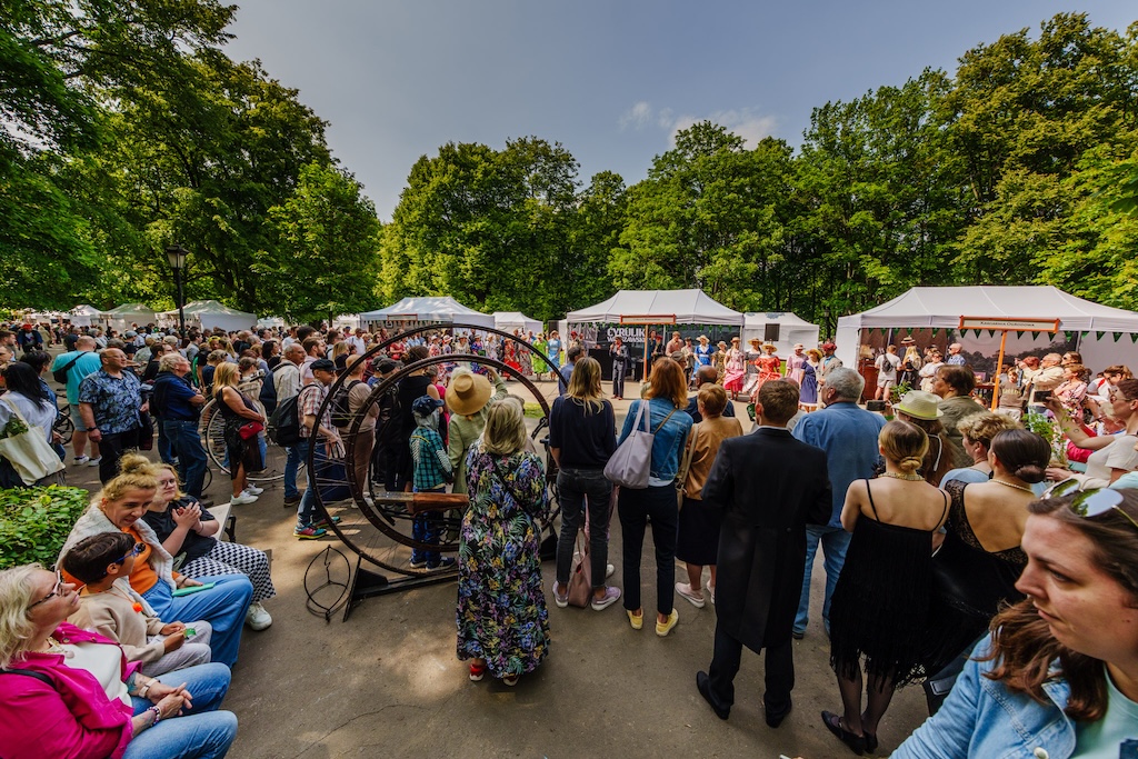Święto Ogrodu Saskiego, czyli urodziny Ogrodu Saskiego. Tłumy widzów stoją w alei parku, oglądając pokaz mody. W tle duże namioty z atrakcjami. Za nimi rosną potężne drzewa.