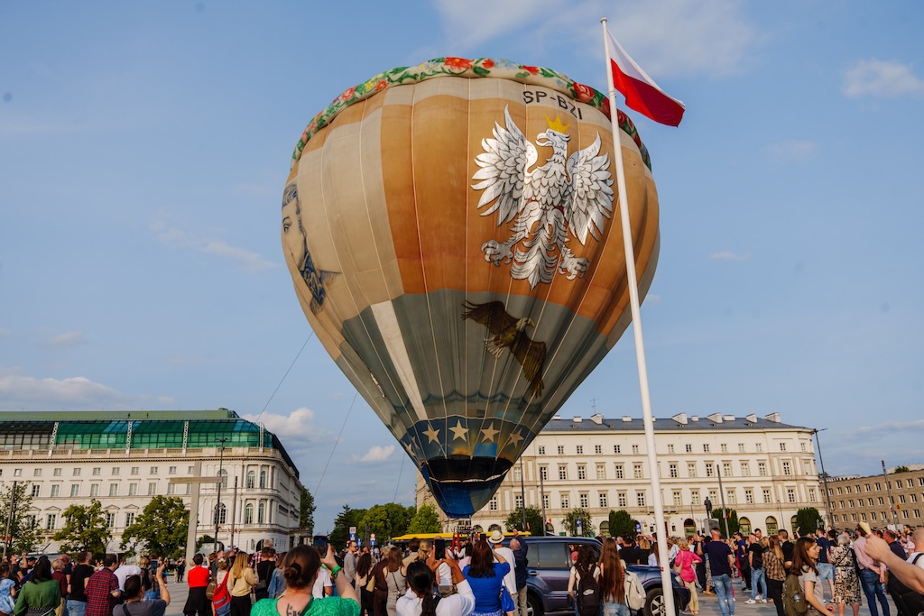 Olbrzymi balon Kościuszko ustawiony na placu Piłsudskiego w Warszawie. Na balonie namalowana jest twarz Kościuszki, polski orzeł i wieniec z kwiatów. Wokół balonu stoi tłum ludzi.