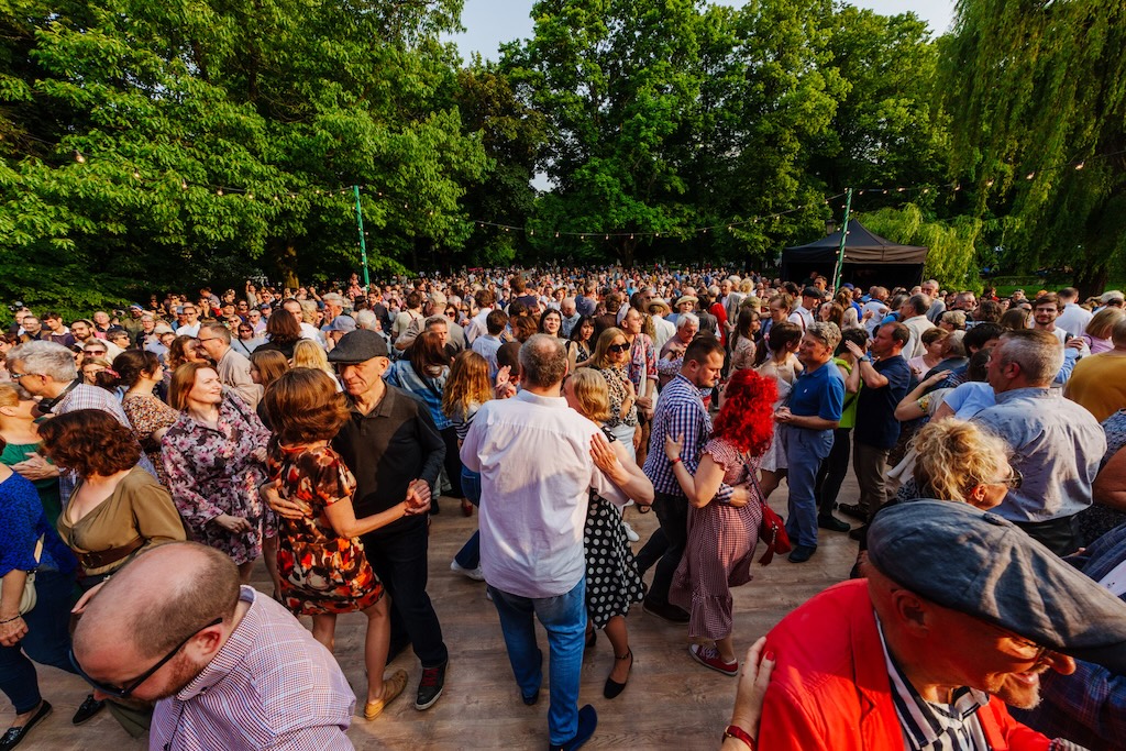 Urodziny Ogrodu Saskiego, czyli doroczne Święto Ogrodu Saskiego. Tłum tańczących ludzi, okolony drzewami. Z przodu tańczą ludzie w parach.