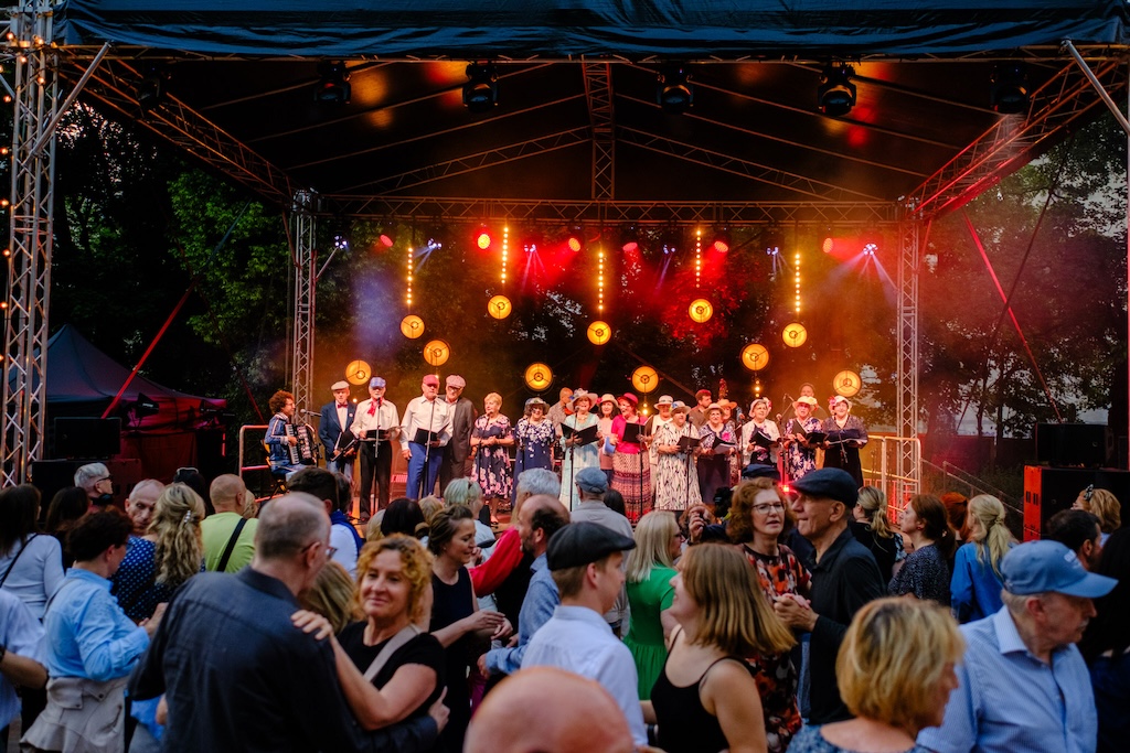 Urodziny Ogrodu Saskiego, czyli Święto Ogrodu Saskiego. Podwyższona scena muzyczna pośród drzew. Na niej chór seniorek w kwiecistych sukienkach i kapeluszach śpiewa do mikrofonów. Przed sceną tańczący tłum.