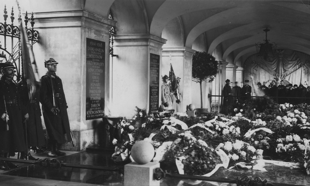 Grób Nieznanego Żołnierza w Warszawie przed wojną. Widok pod kątem, pod arkadami, na płytę grobu, na której ułożono wieńce. Przy płycie stoi warta honorowa. Kawałek dalej, pod arkadami, barierka oddziela tłum ciekawskich. W tle, na końcu arkad, znajduje się ściana z płaskorzeźbą.