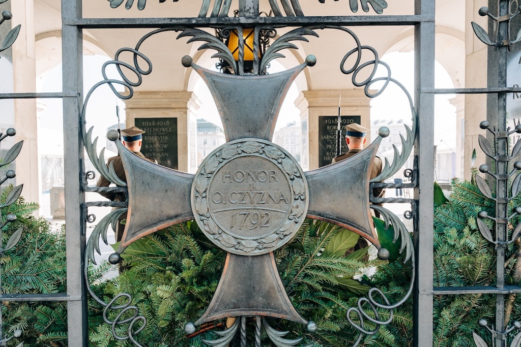 Dekoracja Grobu Nieznanego Żołnierza, czyli krzyż Orderu Viruti Militari na okratowaniu grobu. W tle, za okratowaniem, jest wnętrze grobu i plecy dwóch żołnierzy trzymających wartę honorową.