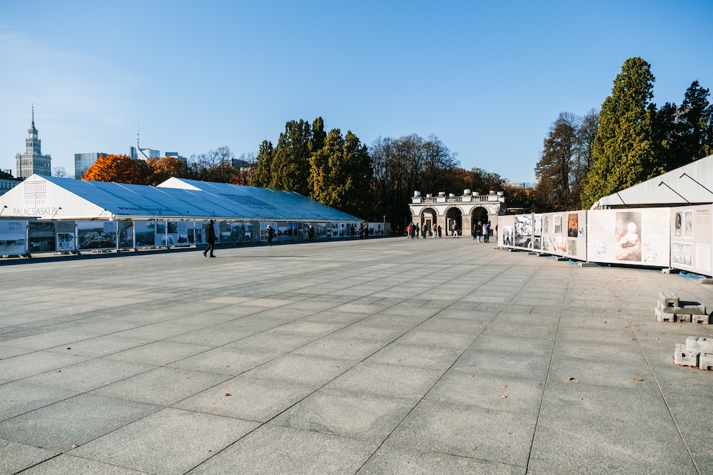 Widok na plac Piłsudskiego w Warszawie. Na pierwszym planie wolna przestrzeń placu, a po bokach ogrodzenie, na którym jest wystawa tablicowa na 100 lat Grobu Nieznanego Żołnierza. W głębi stoi Grób Nieznanego Żołnierza.