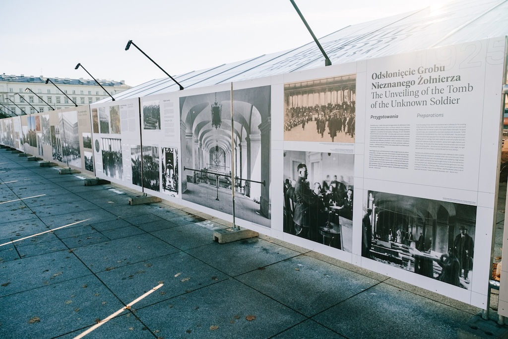 Detal na duże plansze wystawy Wspólne miejsce, wspólna pamięć. 100 lat Grobu Nieznanego Żołnierza w Warszawie na ogrodzeniu na placu Piłsudskiego. Za ogrodzeniem wystaje zadaszenie z logo spółki Pałac Saski.