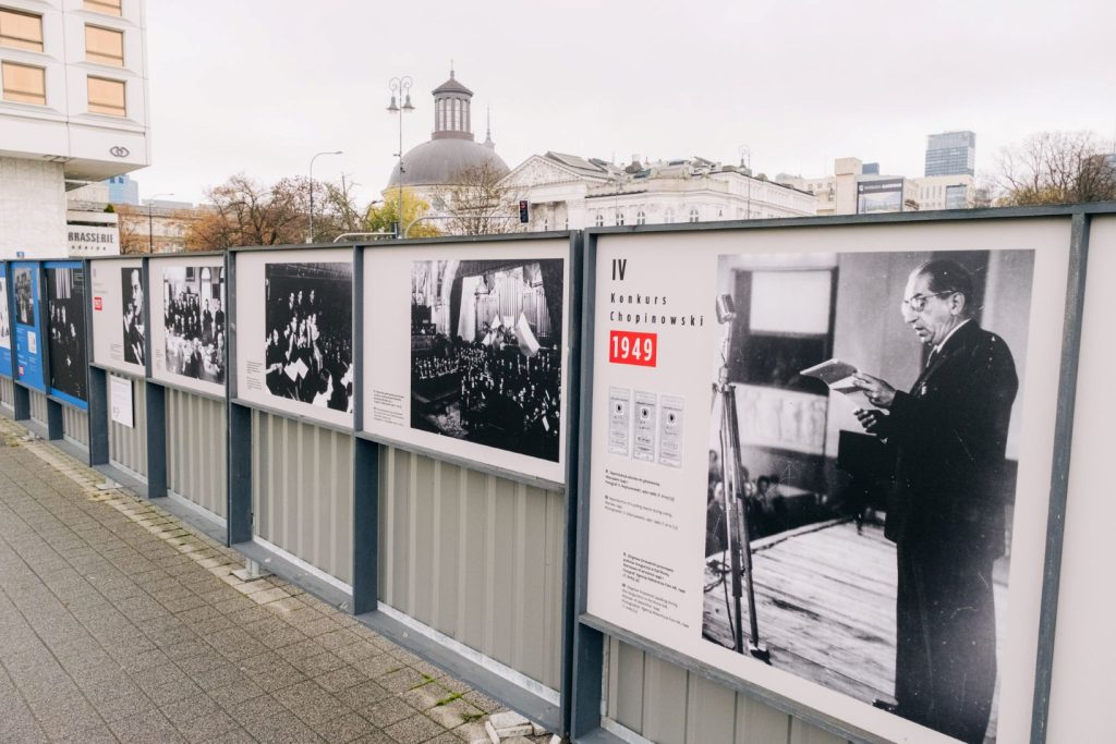 Plansze wystawy zdjęciowej ujęte z ukosa z prawej strony na metalowym ogrodzeniu. Na planszach czarno-białe zdjęcia z Konkursów Chopinowskich, na pierwszym planie po prawej stronie informacje dotyczące IV Konkursu Chopinowskiego w 1949 r. z fotografią wykonaną podczas inauguracyjnego przemówienia Zbigniewa Drzewieckiego w Sali Roma. Na drugim planie, za wystawą, charakterystyczna latarnia warszawska, w tle Zachęta - Narodowa Galeria Sztuki oraz Kościół Ewangelicko-Augsburski Świętej Trójcy. Niebo zachmurzone.