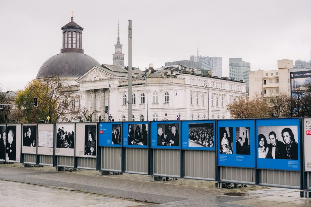 Plansze wystawy zdjęciowej ujęte z ukosa z prawej strony na metalowym ogrodzeniu. Na planszach czarno-białe zdjęcia z Konkursów Chopinowskich. Na drugim planie, za wystawą, charakterystyczna latarnia warszawska, w tle Zachęta - Narodowa Galeria Sztuki, Kościół Ewangelicko-Augsburski Świętej Trójcy, a także fragment Pałacu Kultury i Nauki. Niebo zachmurzone.