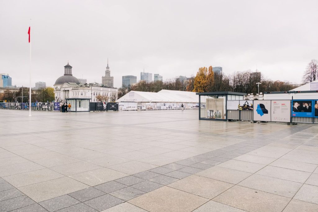 Widok placu marsz. J. Piłsudskiego z umieszczonymi na ogrodzeniu terenu inwestycji odbudowy Pałacu Saskiego planszami dwóch wystaw: "Wspólne miejsce, wspólna pamięć. 100 lat Grobu Nieznanego Żołnierza w Warszawie" oraz "100 lat Konkursu Chopinowskiego w fotografii". Widoczne również ustawione naprzeciw siebie, na dwóch rogach terenu odbudowy przeszklone pawilony. W pawilonie po lewej stronie (w oddali) model 3D odbudowywanych budynków Pałacu Saskiego, Pałacu Brühla oraz kamienic przy ul. Królewskiej 6, 8 i 10/12. Obok pawilonu na wysokim białym maszcie znajduje się flaga Polski. W bliższym pawilonie, po prawej stronie, prezentowane są metalowe tarcze stanowiące niegdyś dekorację Grobu Nieznanego Żołnierza - w tym jedna oryginalna z 1925 r. W tle po lewej Zachęta - Narodowa Galeria Sztuki, Kościół Ewangelicko-Augsburski Świętej Trójcy, Pałac Kultury i Nauki oraz inne wieżowce panoramy Warszawy, po prawej drzewa Ogrodu Saskiego w jesiennej szacie. Niebo pochmurne, płyty placu błyszczące od wilgoci.