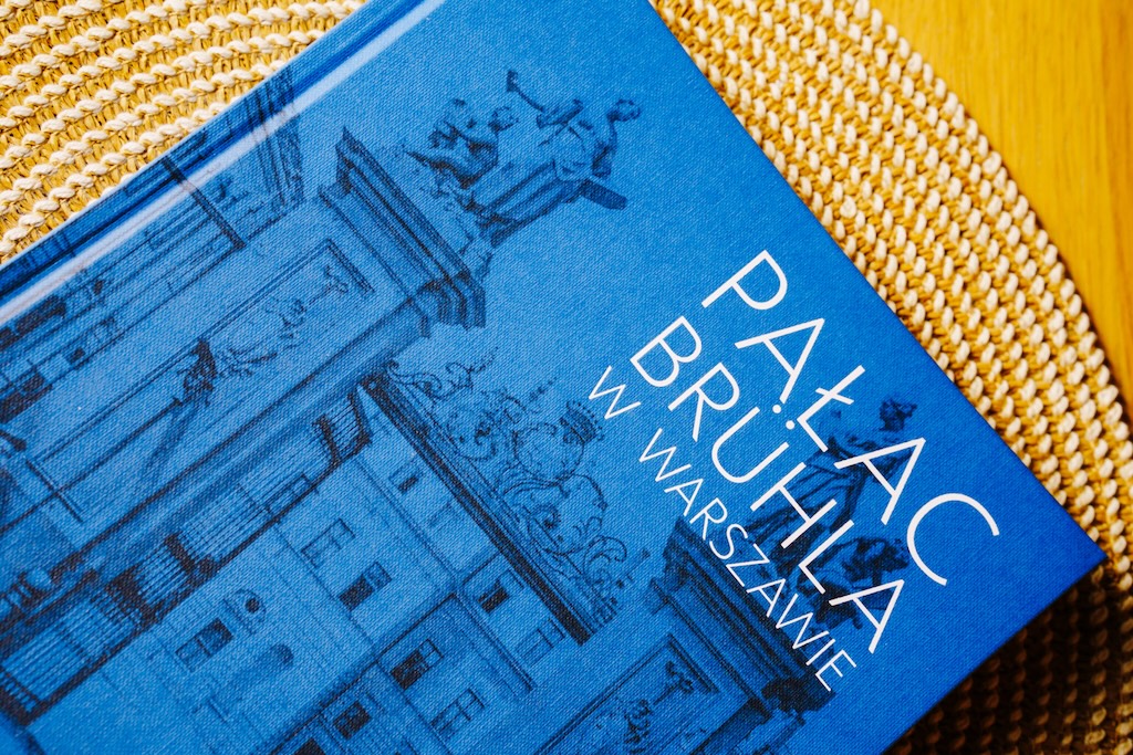 Pałac Brühla w Warszawie, książka w twardej oprawie. Książka leży okładką do góry na ozdobnej, okrągłej macie na drewnianym blacie.