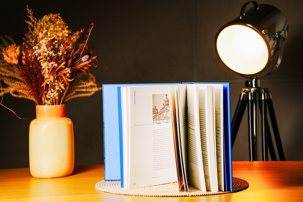 Pałac Brühla w Warszawie, otwarta książka postawiona na ozdobnej, okrągłej macie na drewnianym blacie. Książka jest postawiona stronami do odbiorcy, by móc zobaczyć jej wnętrze. Za książką, po lewej stoi ceramiczny wazon z bukietem suszonych roślin. Po prawej, zza stołu wyłania się lampa na trójnogim stojaku.