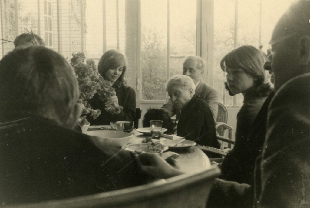 Czarno-białe archiwalne zdjęcie grupy osób (od lewej: Barbara Kwiatkowska, Maria Dąbrowska, za nią Jerzy Giedroyc, Tula Kowalska, Jerzy Andrzejewski) siedzących przy stole w jasnym pomieszczeniu w domu "Kultury" paryskiej. Na stole stoją filiżanki, talerze i bukiet kwiatów. W tle za oknami widoczne rośliny ogrodowe.