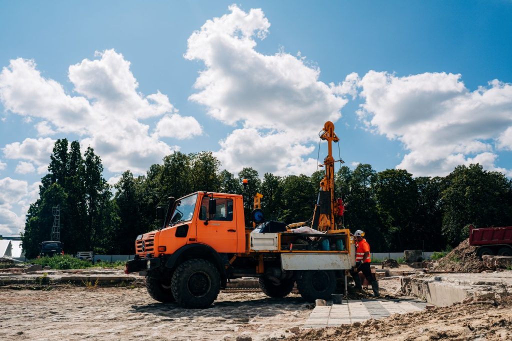 Pomarańczowy samochód ciężarowy z wiertnicą i pracownikiem na piaszczystym placu odbudowy Pałacu Brühla w Warszawie, na tle ciemnozielonych drzew, błękitnego nieba i białych chmur.