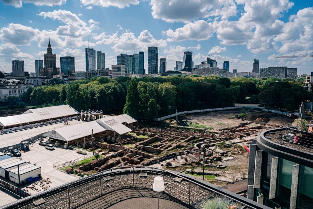 Widok z góry na fragment tereny odbudowy zachodniej pierzei placu Piłsudskiego, z odsłoniętymi reliktami piwnic Pałaców Saskiego i Brühla oraz panoramą Warszawy pod błękitnym niebem z białymi chmurami.