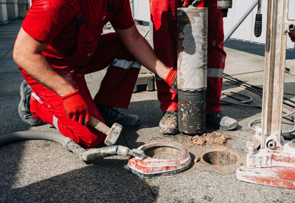 Dwóch pracowników w czerwonych strojach przy pobieraniu cylindrycznej próbki podczas badań geologicznych na placu odbudowy Pałaców Saskiego i Brühla w Warszawie.  