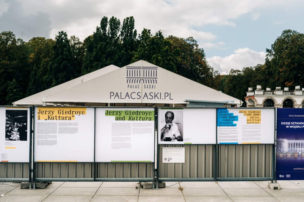 Plenerowa wystawa „Jerzy Giedroyc i «Kultura»” na planszach na ogrodzeniu terenu odbudowy Pałacu Saskiego ustawionym przed białym zadaszeniem namiotowym z napisem PALACSASKI.PL. Na prawo od zadaszenia fragment Grobu Nieznanego Żołnierza. Na jednej z plansz widoczny jest portret Jerzego Giedroycia.