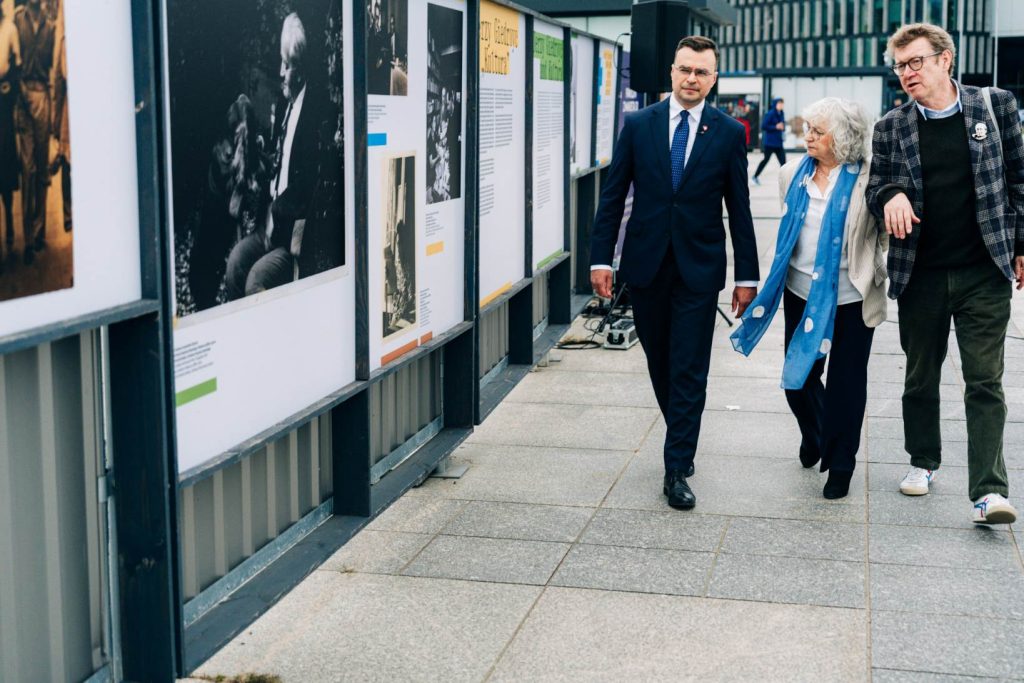 Trzy osoby (od lewej: Maciej Wróbel, Anna Bernhardt i Stanisław Mancewicz) przechodzą obok plansz plenerowej wystawy „2026: Rok Jerzego Giedroycia – w 120. rocznicę urodzin”.